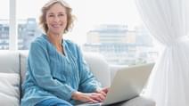 Content blonde woman sitting on her couch using laptop smiling at camera at home in the sitting room