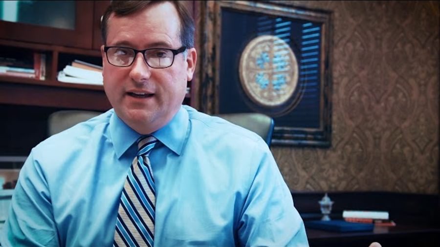 A man in a blue shirt and tie looking into the camera at his desk.