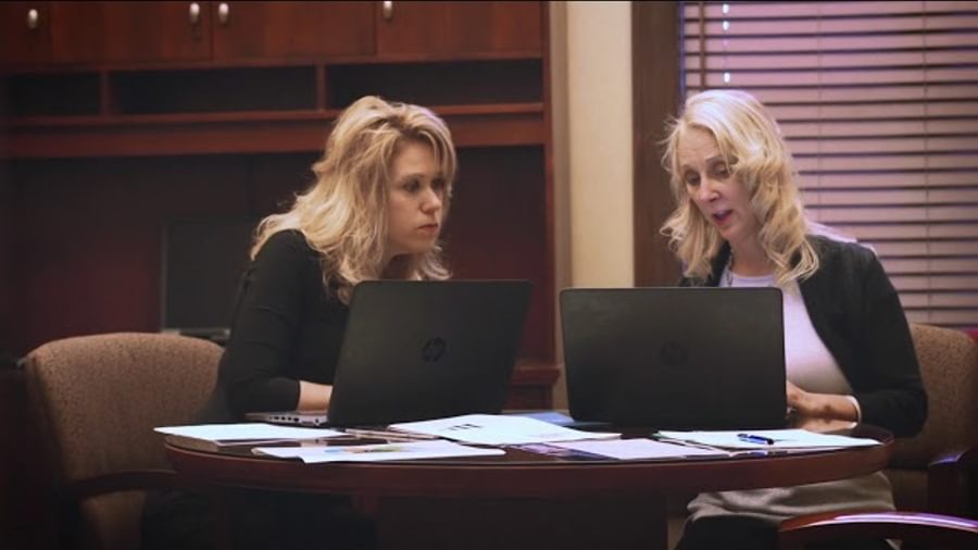 Two women at a desk. They are wearing business attire, looking at a computer and talking.