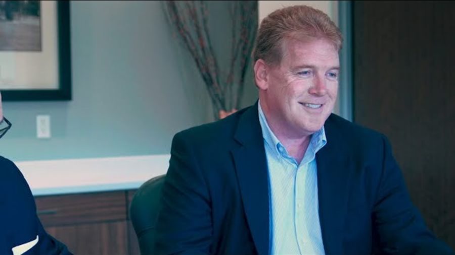 A man in business attire at a desk. He is not wearing a tie, and is smiling in confidence.