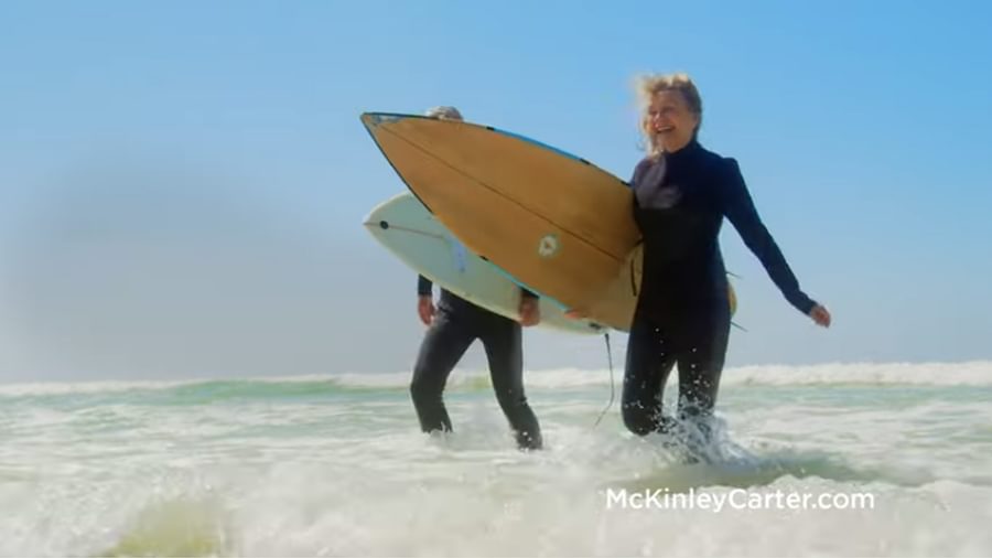 A middle-aged woman, walking out of the ocean holding a surf board.